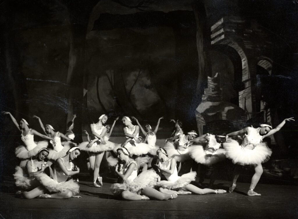 Valkoisiin tutuihin ja päähineisiin pukeutuneita balettitanssijoita mustavalkoisessa kuvassa. Tanssijat ovat asettuneet kuvan keskelle muodostelmaan, jossa osa tanssijoista seisoo ja osa istuu. Heidän taustallaan on tummia lavasteita.