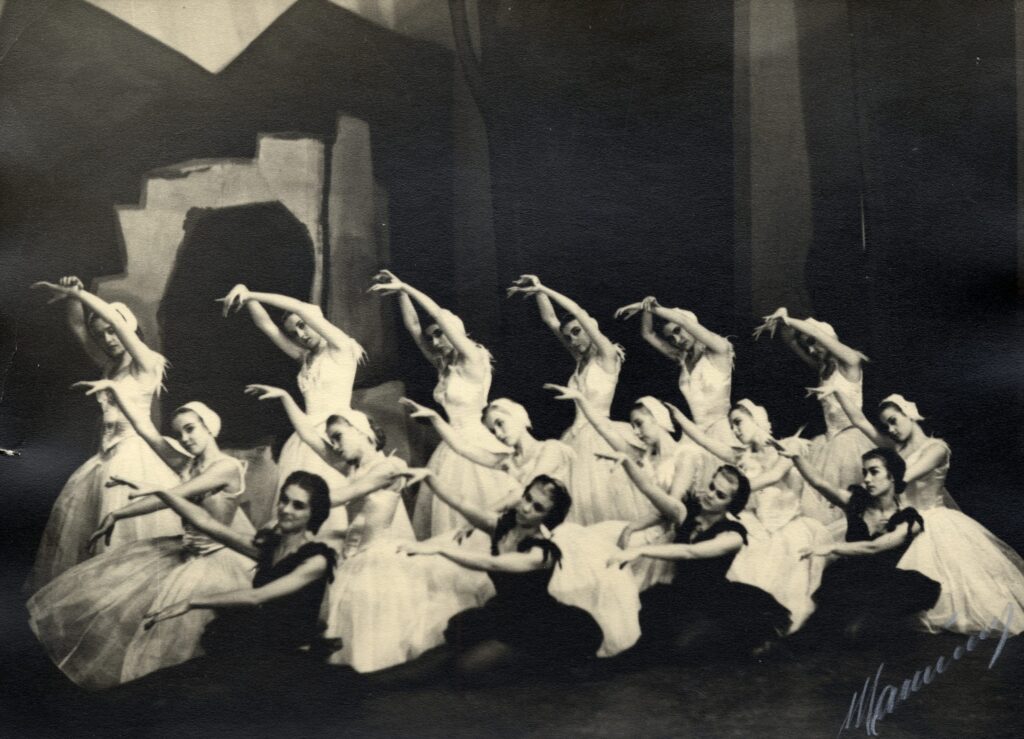 Mustavalkoinen kuva, jossa 16 naispuolista balettitanssijaa kolmessa rivissä. Etummaisilla tanssijoilla on mustat asut, lopuilla valkoiset. Osa tanssijoista istuu ja osa on polviensa varassa.