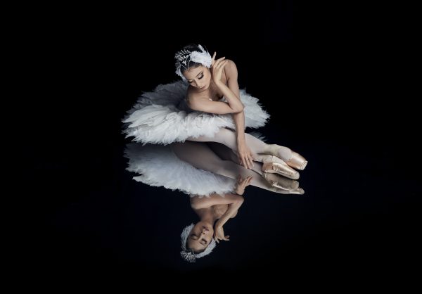 Vaalea-asuinen balettitanssija istuu ja katsoo tummaan lattiaan, josta näkyy hänen heijastuksensa. 