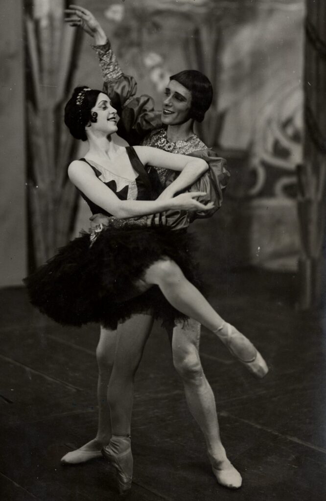 Kaksi balettitanssijaa msutavalkoisessa kuvassa. Naistanssijalla on musta tutu ja kärkitossut. Puhvihihaiseen asuun pukeutunut miestanssija tukee häntä pitämällä kättä vyötäisillä.  