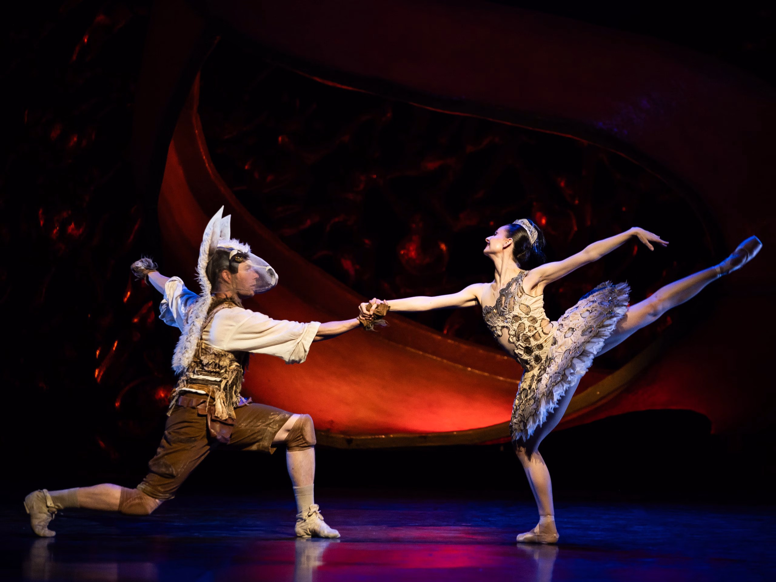 Aasinpäinen miestanssija polvistuneessa asennossa ja tutuasuinen naistanssija arabeskissa tarttuvat toisiaan kädestä tanssiessaan lavalla.