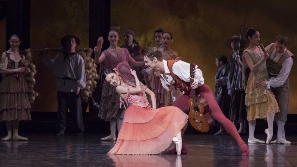 Persikanväriseen röyhelömekkoon puettu nainen on polvellaan toinen jalka eteen koukistettuna ja piiloutuu viuhkan taakse. Punaisiin trikoisiin ja valkoiseen paitaan puettu mies kurottautuu hymyillen häntä kohti. Taustalla on ihmisiä hämärässä valaistuksessa.