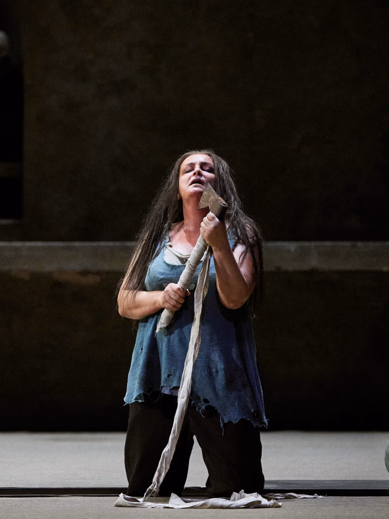 Nainen seisoo polvillaan ja pitelee kirvestä. Hänen silmänsä ovat kiinni ja suu auki. Hänellä on pitkä tumma tukka ja päällään sininen, rikkinäinen hihaton paita ja tummat housut. Tausta on hämärä.